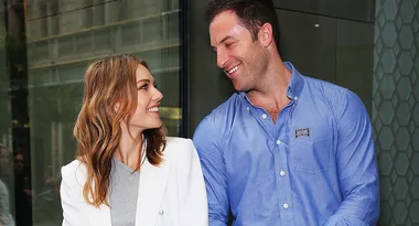A smiling woman in a white blazer looks at a man in a blue shirt. They stand outdoors near a glass building.