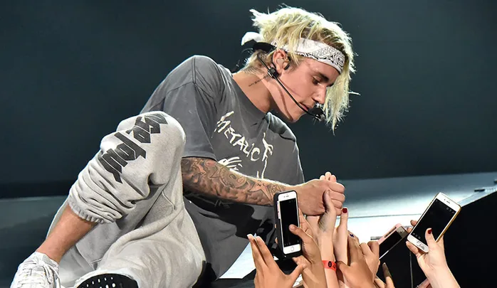 Musician wearing a bandana interacts with fans, holding hands from stage, surrounded by raised phones capturing the moment.