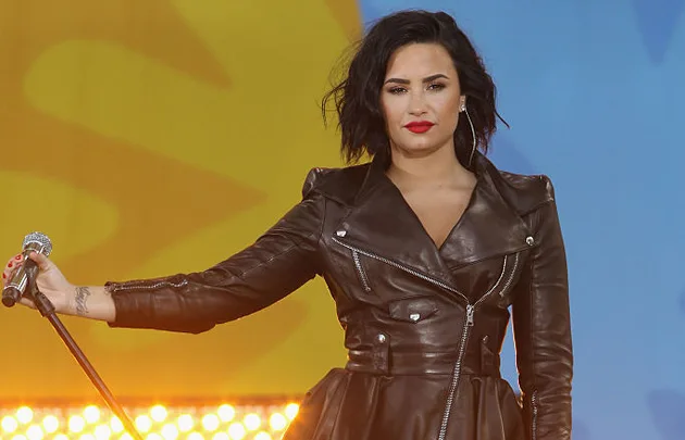 Singer in a black leather jacket holds a microphone on stage with a colorful background.
