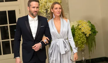 Couple dressed formally, man in a black suit with bowtie, woman in a silver gown, posing arm in arm indoors.