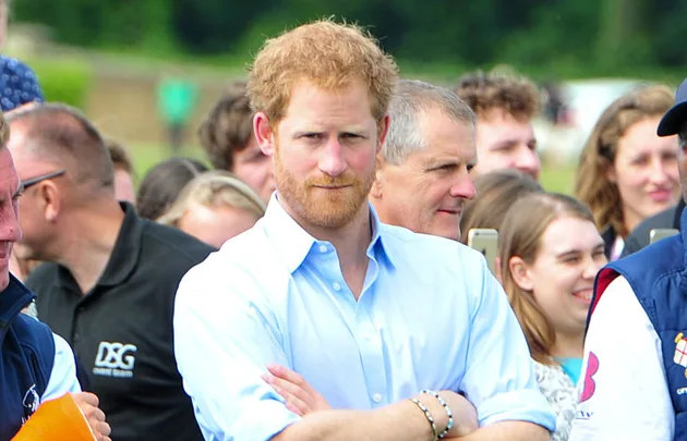 A man with a beard in a light blue shirt stands in a crowd outdoors, crossing his arms and looking forward.