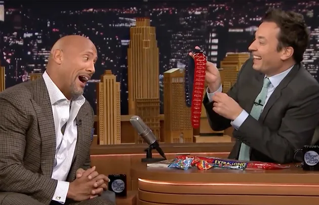 Two men on a talk show desk, one holding a large red candy, both laughing, cityscape backdrop.