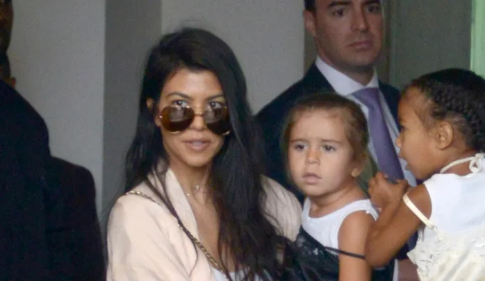 Woman wearing sunglasses holds a child, with another child nearby and a man in the background.
