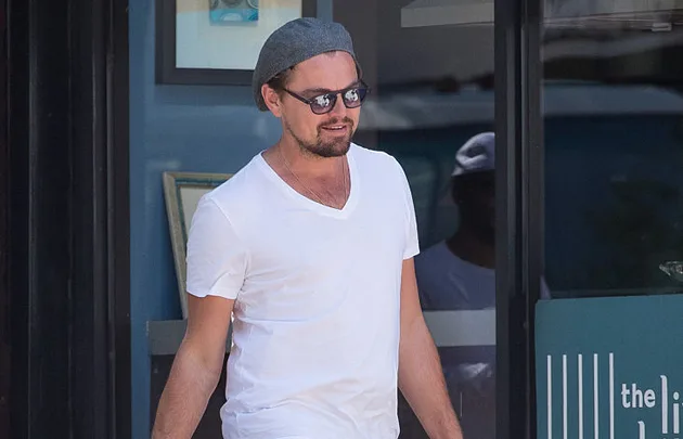 Man in sunglasses and a grey hat, wearing a white t-shirt, walking outside a glass door.