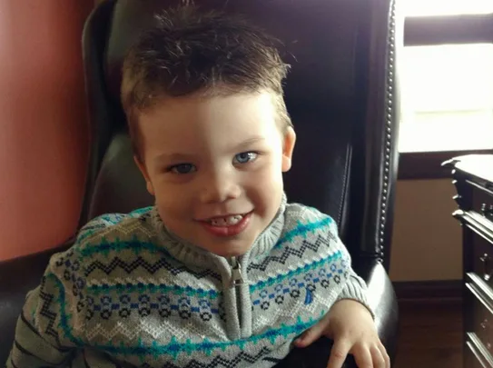 A young boy with a patterned sweater sits on a leather chair, smiling at the camera.