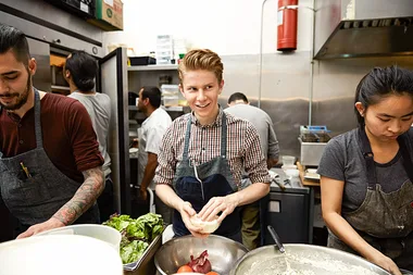 Cooks in a busy restaurant kitchen, with one person smiling and preparing food while others work around him.
