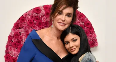 Two people posed in front of a pink rose wall, one in a blue dress and the other in a denim jacket, smiling together.