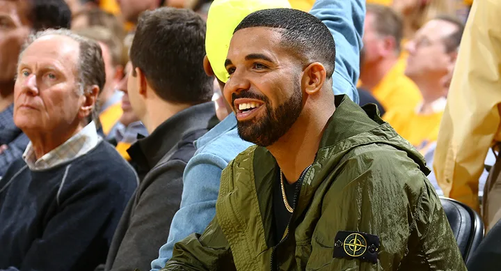 A smiling man in a green jacket sits among a crowd at a sports event.