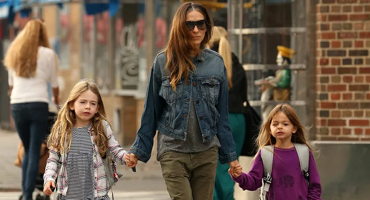 A woman in sunglasses walks holding hands with two young girls on a city street.
