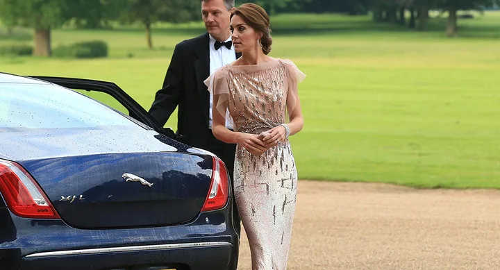 A woman in an elegant gown stands beside a black Jaguar, with a man in a suit in the background.