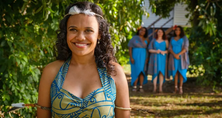 A smiling woman in a patterned dress stands in a garden, with three women in blue dresses blurred in the background.