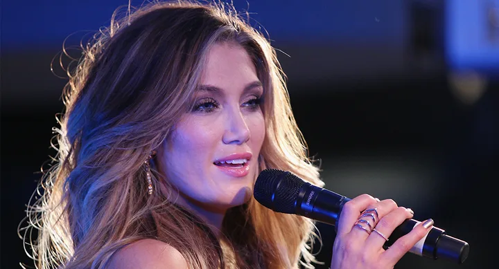A woman with long hair sings into a microphone on stage, illuminated by blue lighting.