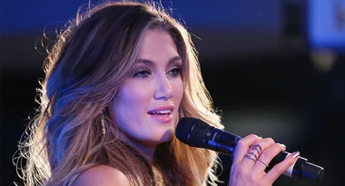 A woman with long hair sings into a microphone on stage, illuminated by blue lighting.