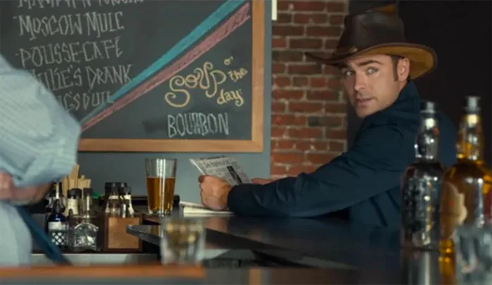 Cowboy-hat man at bar reads menu; chalkboard says "Soup of the day: Bourbon"; bottles and glass on counter.