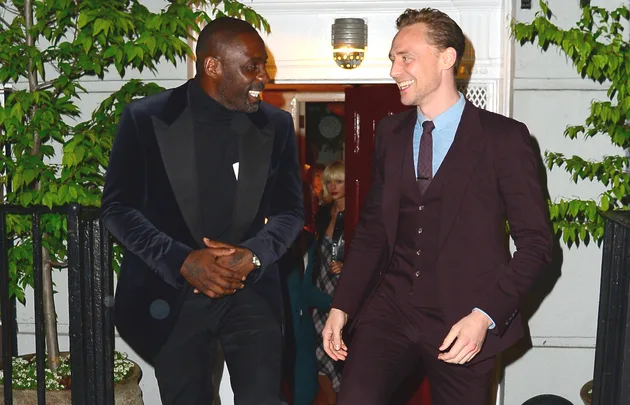 Two men in suits smiling and talking outside a building with greenery around.