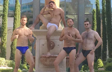 Four men in swim briefs pose in front of a fountain outdoors.