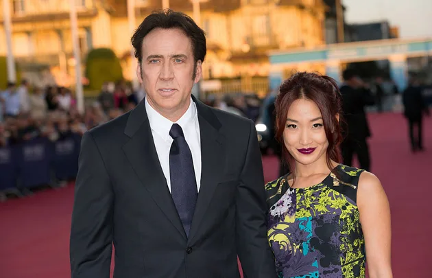 A man in a suit and a woman in a floral dress on a red carpet with a blurred crowd in the background.