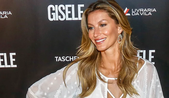 Woman in white dress posing and smiling at an event with "GISELE" text in the background.