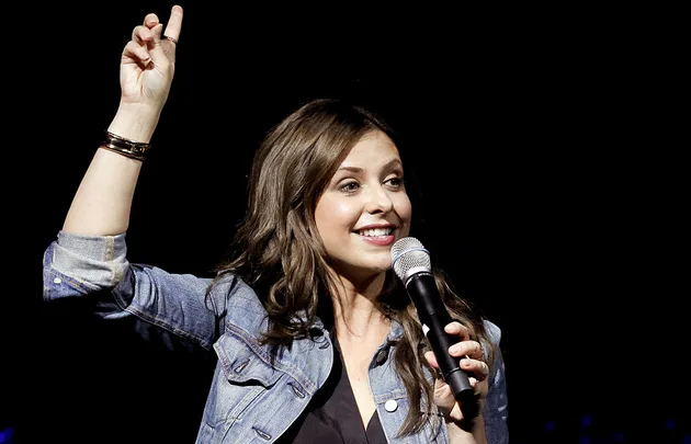 Woman holding a microphone on stage, smiling and raising her hand, wearing a denim jacket.