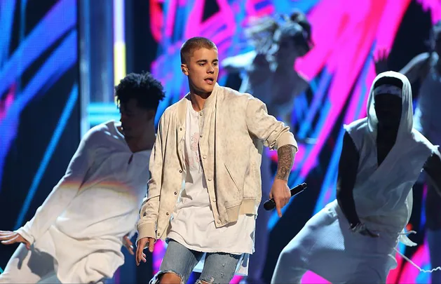 Singer performs on stage with dancers, wearing a beige jacket and holding a microphone, colorful background.