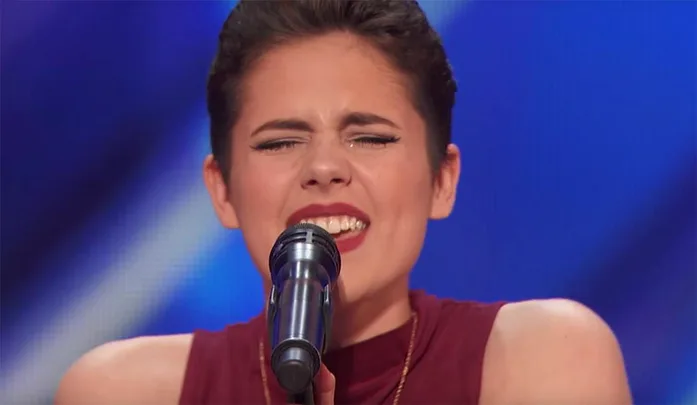 A person passionately singing into a microphone on stage, eyes closed, wearing a sleeveless red outfit.
