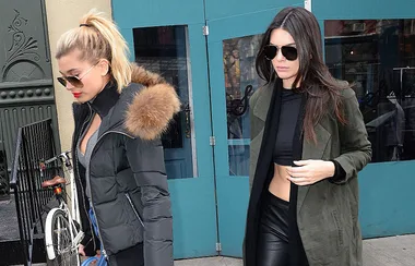 Two women in winter jackets and sunglasses walk past a turquoise door on a city street.