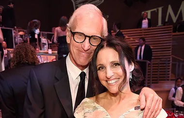 Man in glasses and black suit embracing woman in white dress, both smiling at an event.