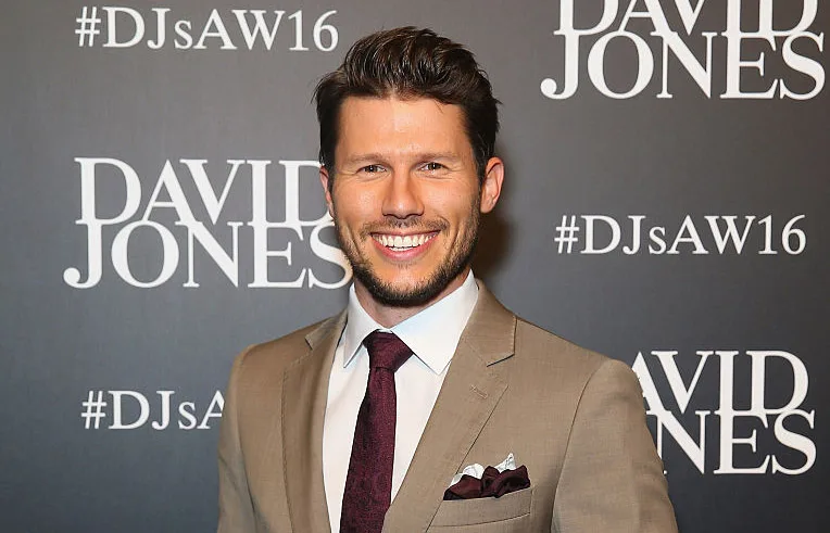 A man in a tan suit and maroon tie smiles at a David Jones event backdrop.