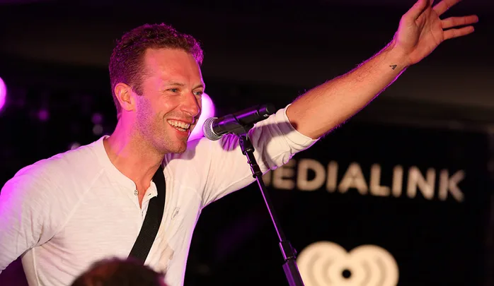 Singer in white shirt performing on stage, smiling, and reaching out to audience. Microphone positioned in front.