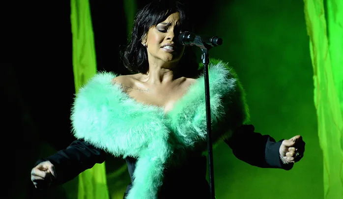 Rihanna performing on stage in a green fur shawl, with eyes closed and hands raised, under green lighting.