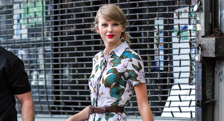 A woman in a floral dress and red lipstick walks past a metal gate, accompanied by a person in black.