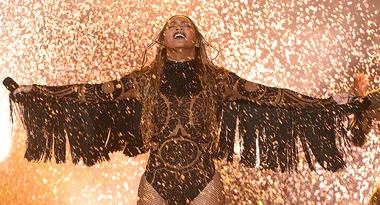 Performer in black costume on stage with arms outstretched, surrounded by golden sparks, conveying intense emotion.