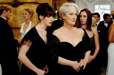 Three women in elegant black dresses at a formal event, with people mingling in the background.