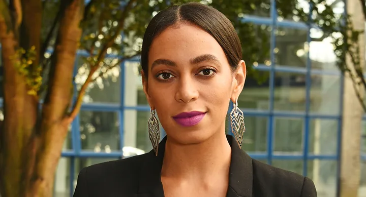 Portrait of a person with purple lipstick, wearing a black blazer and statement earrings, outdoors with trees and windows.