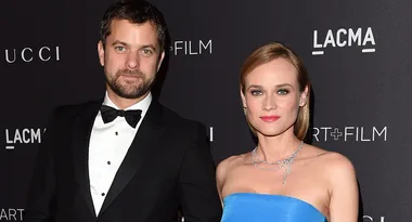 Two people posing elegantly on a red carpet at a LACMA event. The woman wears a blue dress, the man a black tuxedo.