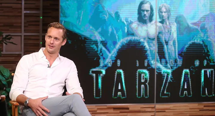 Actor in a white shirt sits in front of a "The Legend of Tarzan" movie poster.