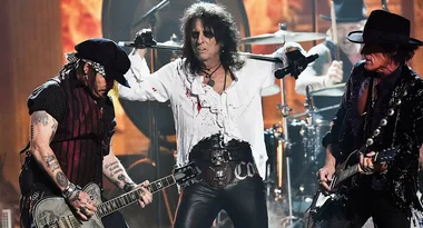 Rock band performing on stage with two guitarists and a vocalist in theatrical attire.