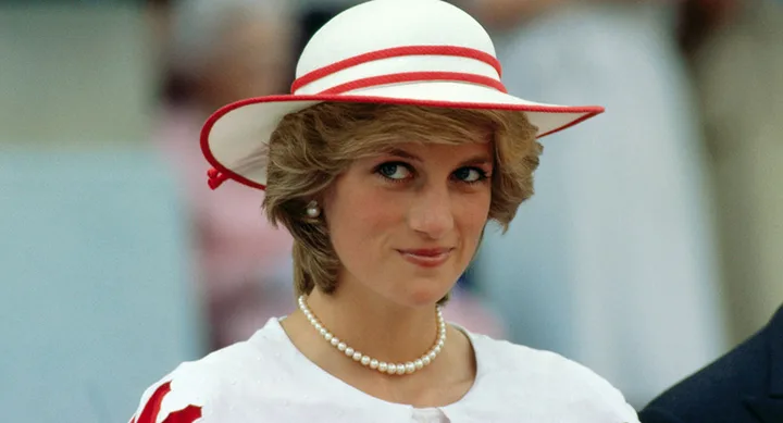 A woman in a white hat with red trim, wearing pearl earrings and necklace, smiling slightly.