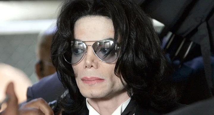 A person with long dark hair wearing sunglasses and a white shirt under a dark coat, indoors.
