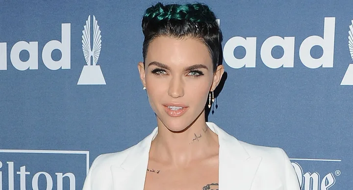 A person with dark hair styled in braids, wearing a white suit, poses at a GLAAD event.