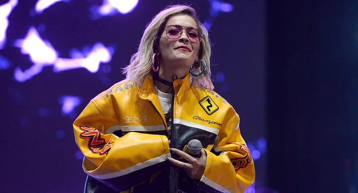 Singer wearing a yellow racing jacket, holding a microphone, performs on stage with purple lights in the background.
