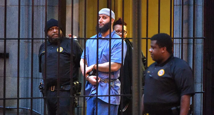 Man in blue prisoner attire escorted by security guards outside a building behind bars.