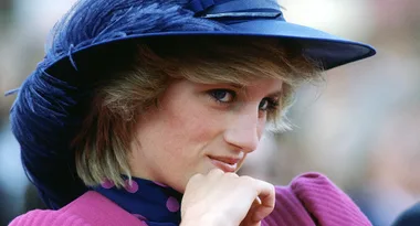 A woman in a purple outfit and blue hat gazes thoughtfully to the side, with a soft, natural background.