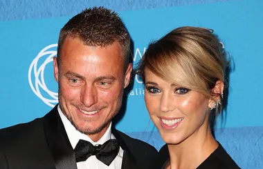 A couple poses at a formal event, both smiling; the man in a tuxedo, the woman in a black dress with diamond earrings.