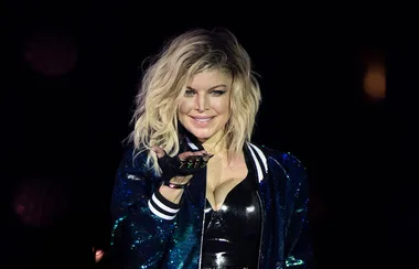 A performer with wavy blonde hair blows a kiss to the audience, wearing a shiny blue jacket and a black top.