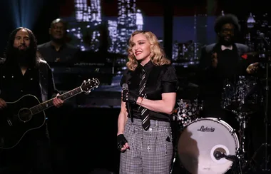Singer performs on a stage with musicians, wearing black top and checkered pants, holding a microphone.