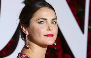 I don't know who this person is, but they have dark hair, are wearing a red dress, and long earrings, on a red floral background.