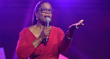 Person in red dress holding a microphone on stage with a purple-lit background.