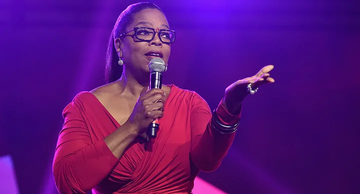 Person in red dress holding a microphone on stage with a purple-lit background.