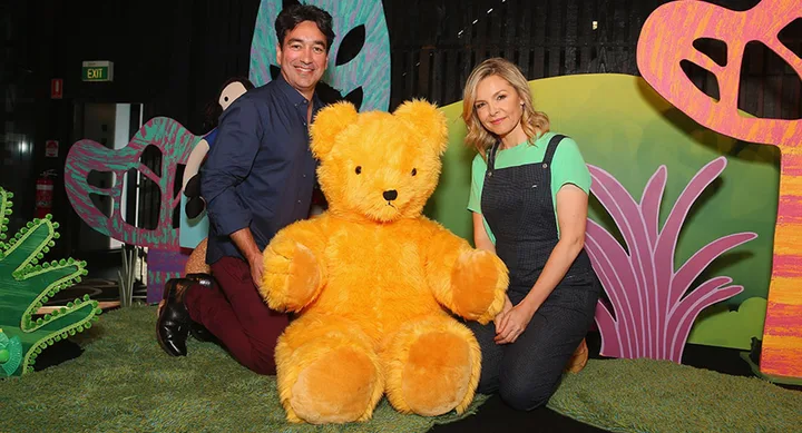 Two people kneel beside a large yellow teddy bear in a brightly colored, whimsical setting.